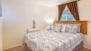 A bedroom with a wooden headboard, a patterned bedspread, a bedside table with a lamp, and a window with brown curtains. - Crabbin Cabin Westport (Westport)