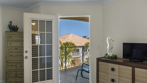 Quarto com porta de vidro deslizante para uma varanda com vista para uma casa e palmeiras. Há uma televisão em um armário de madeira e um pavão decorativo na parede.