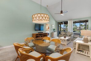 Ein modernes Wohnzimmer mit einem Esstisch aus Glas, umgeben von Korbstühlen, einem Deckenventilator und einem großen Fenster mit Blick auf Palmen.