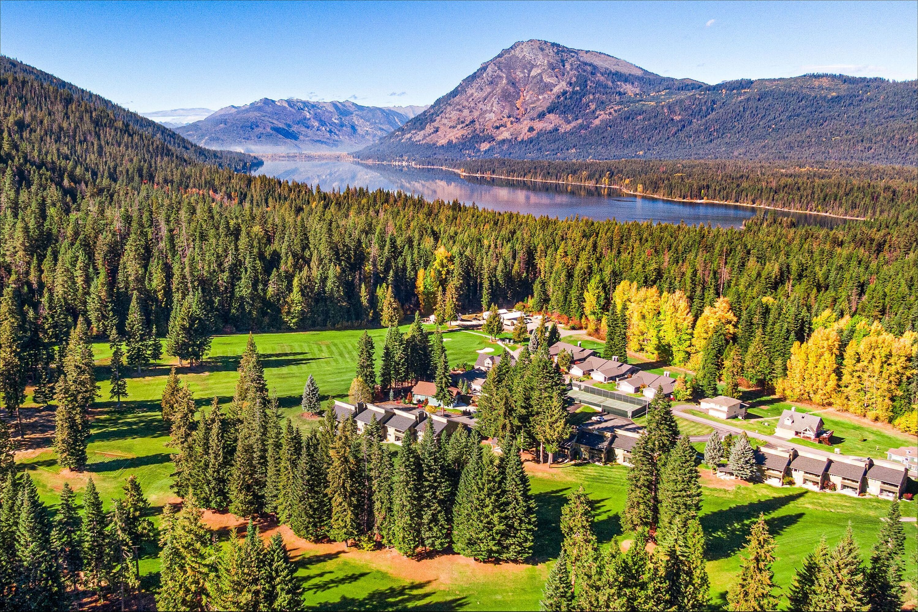 A resort with multiple buildings, a golf course, and a lake surrounded by forested mountains.