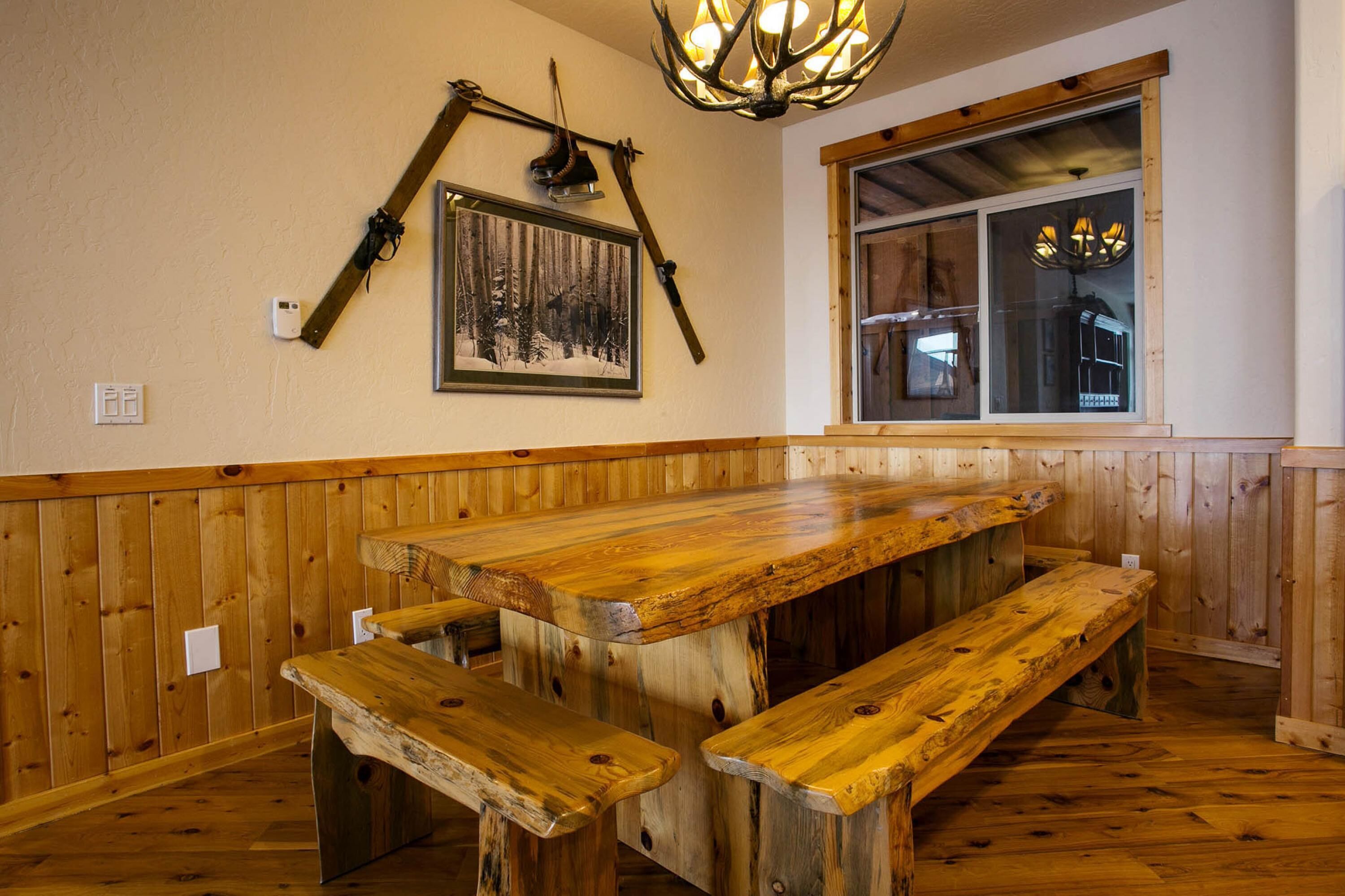 A rustic wooden interior with a large wooden table and benches, a framed forest photograph on the wall, and a window with a view of the outside.