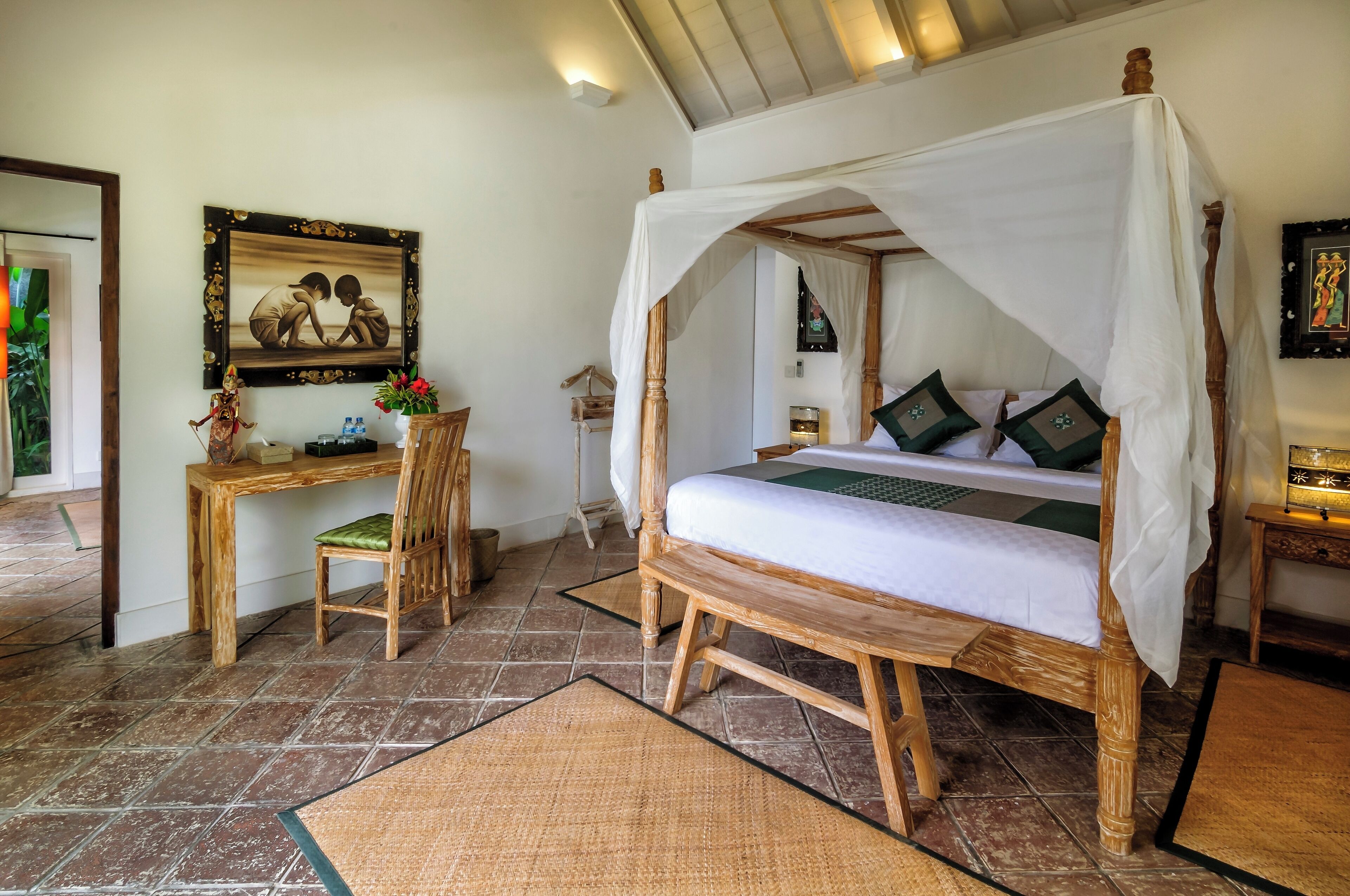 A room with a four-poster bed, a wooden desk with a chair, a framed picture on the wall, and a small table with a vase.