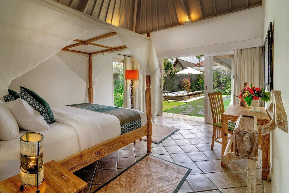Un dormitorio con cama con dosel, una mesita de noche con lámpara, y vista al jardín a través de una puerta de cristal.