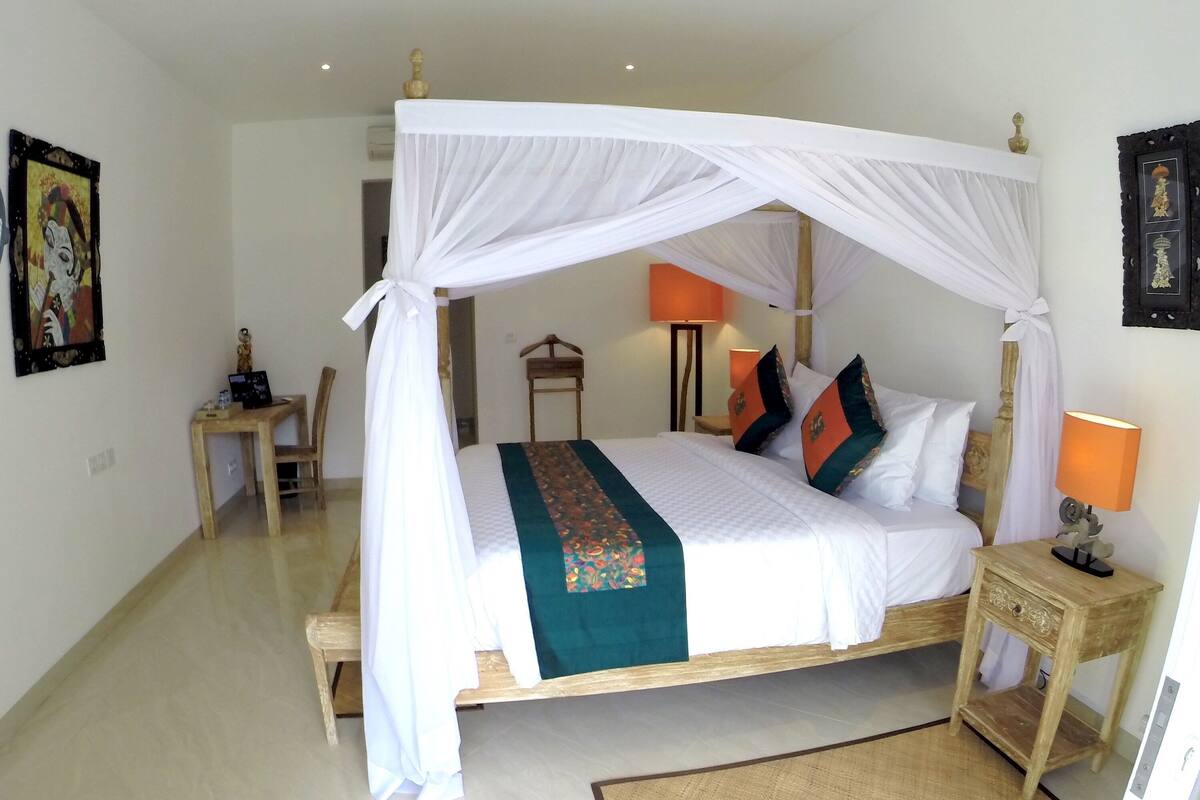 Un dormitorio ordenado con cama con dosel, una mesita de noche con lámpara, una silla y un escritorio con ordenador portátil.