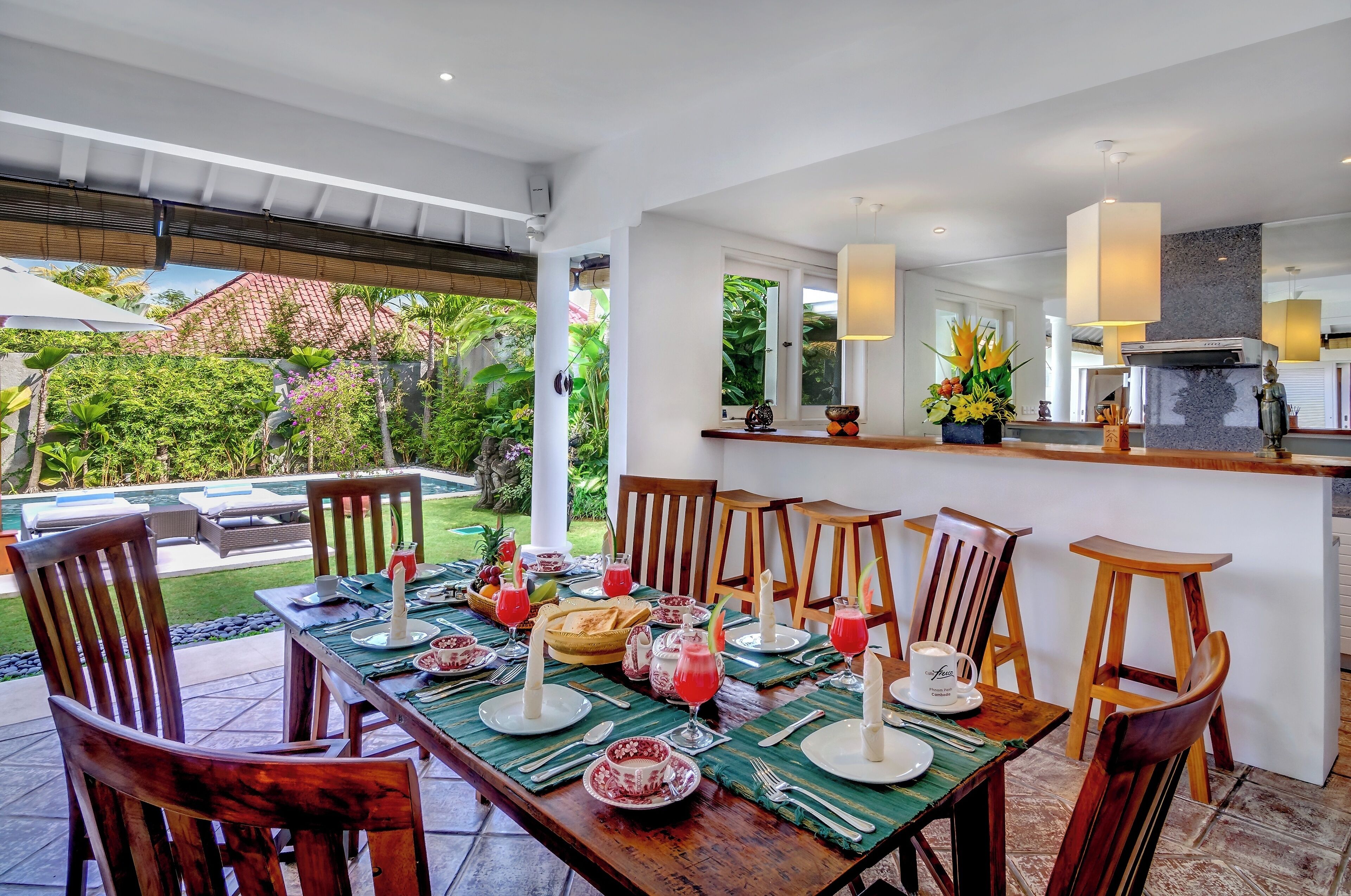 Uma sala de jantar com vista para a piscina, cadeiras de madeira, uma mesa longa posta para refeição e uma área de cozinha com balcão e luminárias pendentes.