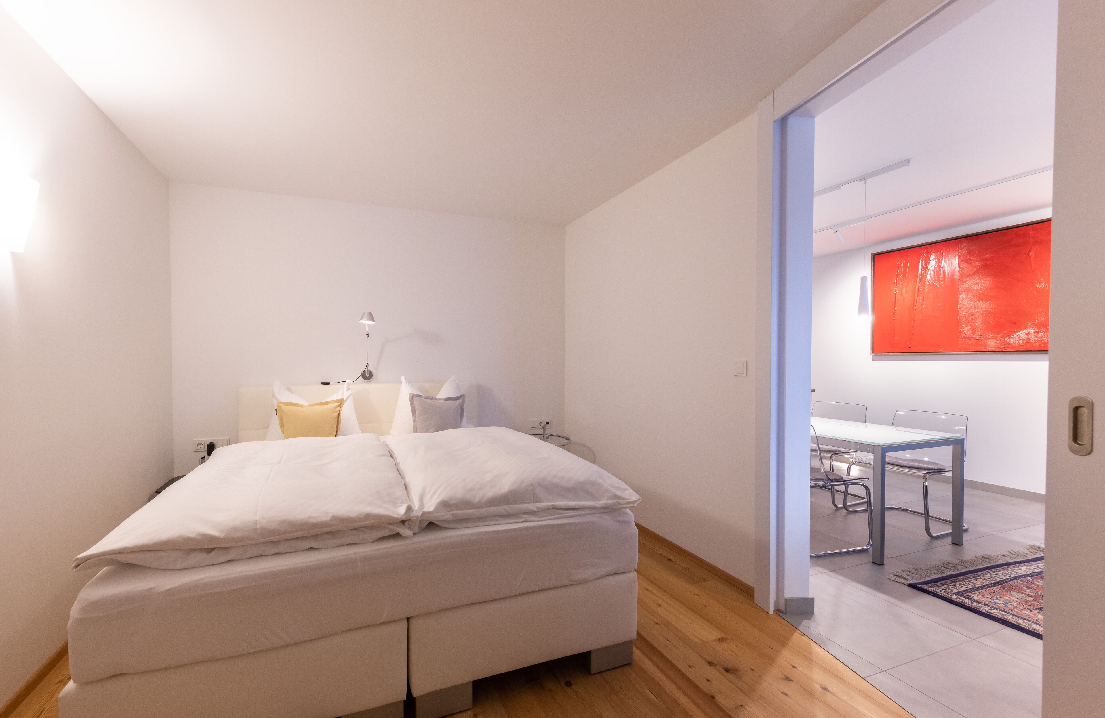 A bedroom with a bed, bedside tables, and a view into a dining area with a table and chairs.