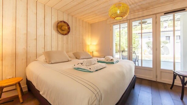 A hotel room with a wooden ceiling, a large bed with white linens, a bedside table, a chair, and a view through a glass door.