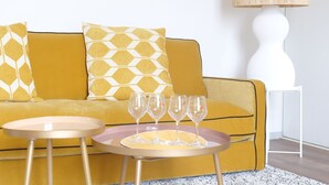 Un sofá amarillo con cojines estampados, una mesita redonda con una bandeja que sostiene cuatro copas de vino y una lámpara blanca sobre una mesita auxiliar blanca.