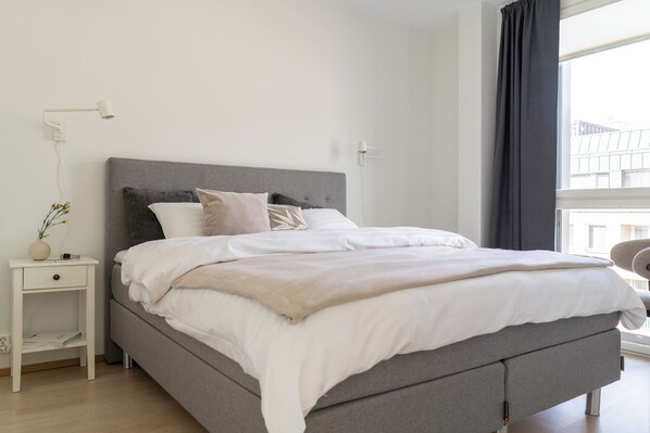 A modern bedroom with a grey upholstered bed, white bedding, a bedside table with a lamp, and a window with a view of buildings. - Modern Haven in Downtown Tampere (Tampere)