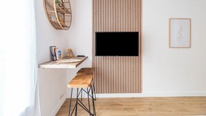 Una habitación moderna con suelo de madera, pared blanca, televisor montado, panel de pared de listones de madera, una silla de mimbre e imagen enmarcada.