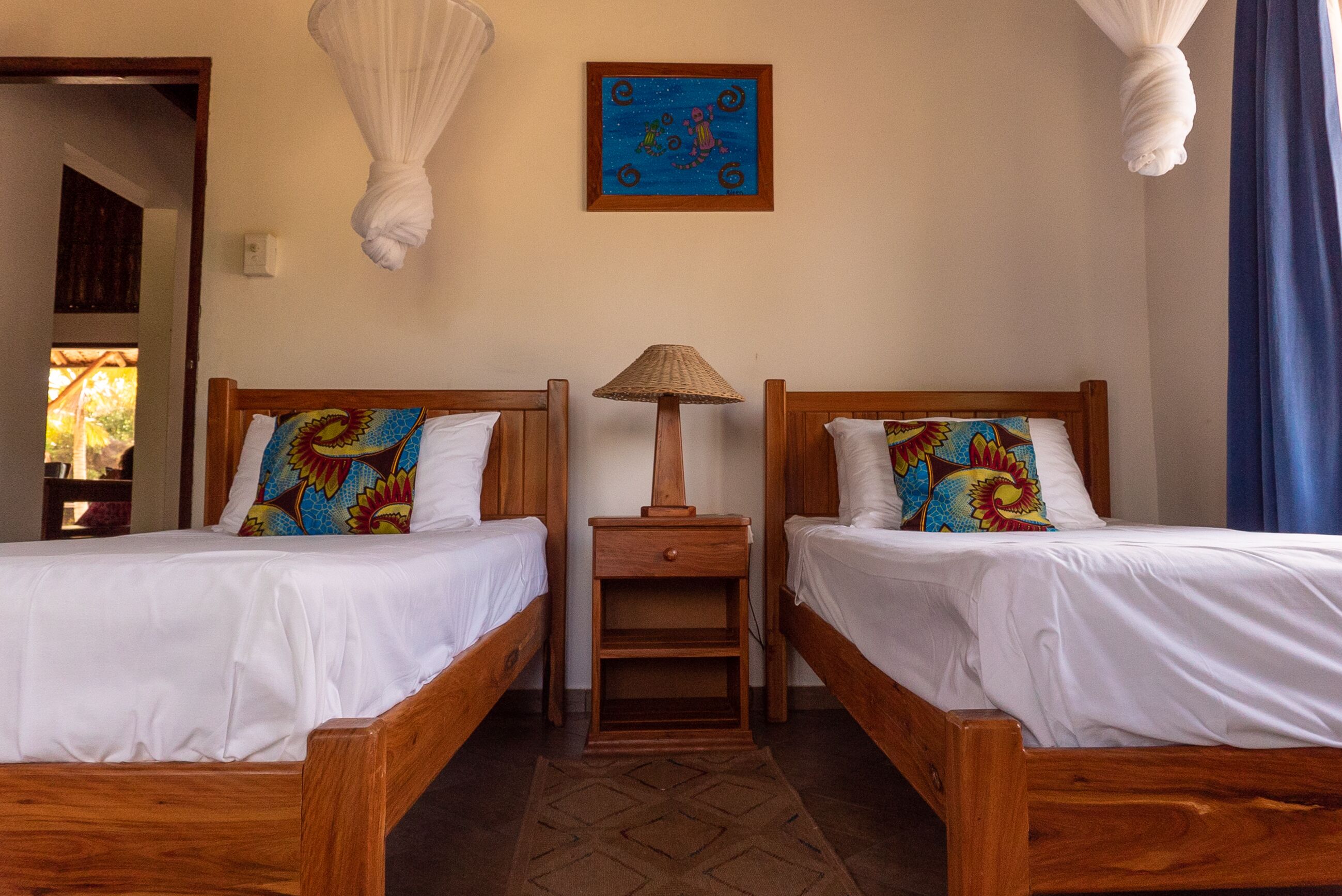 Una habitación con dos camas de madera, cada una con almohadas de colores y una mesita de noche de madera entre ellas. Se observa una lámpara colgante sobre las camas.