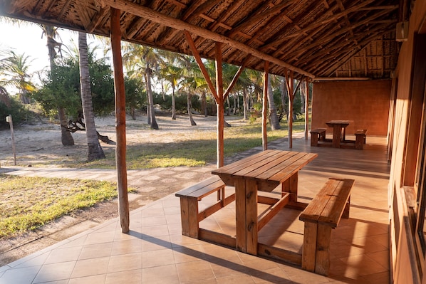 A covered outdoor area with a wooden table and benches, surrounded by palm trees and a sandy ground. - Morrumbene Beach Resort (Morrumbene)