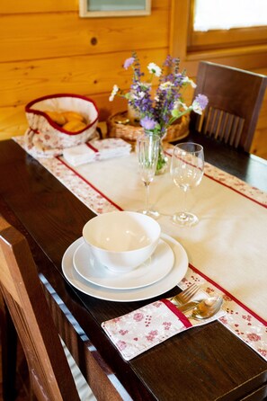 A wooden dining table set with white plates, bowls, and cutlery, accompanied by a vase of flowers and two wine glasses.