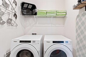 Vaskerum med to Whirlpool vaskemaskiner, en sort kurv og en hylde med foldede håndklæder.