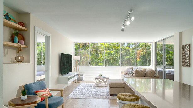 A modern living room with a sofa, chairs, a coffee table, and a TV. There are shelves with decorative items and a large window with a view of greenery. - Spacious Remodeled Across from Kamaole 3 Beach (Kihei)