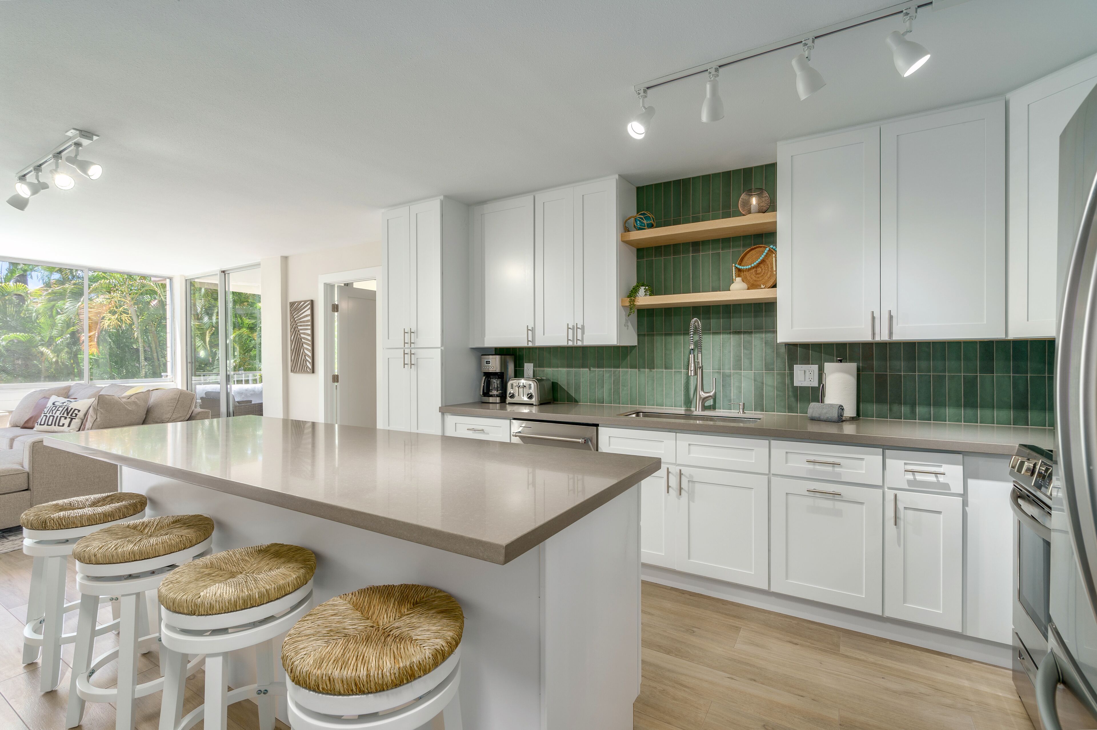 A modern kitchen with white cabinets, a large island with stools, a green backsplash, and a view of the outdoors.