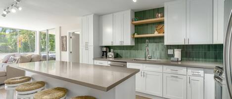 A modern kitchen with white cabinets, a large island with stools, a green backsplash, and a view of the outdoors.