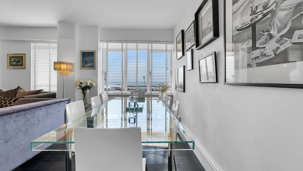 A modern living room with a glass dining table, white sofas, and a large window with blinds. - Deco Seafront - Seafront Apartment - Sleeps up to 6 (Brighton)