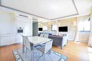 A modern living room with a sofa, a dining table with chairs, a flat-screen TV mounted on the wall, and a kitchen area with cabinets and appliances.