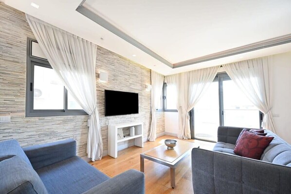 A modern living room with a flat-screen TV, grey sofas, a coffee table, and large windows with curtains.