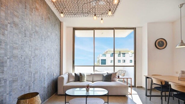 A modern living room with a sofa, a round coffee table, a dining table with chairs, and a large window offering a view of buildings.