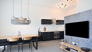 A modern kitchen with a dining area, featuring a large flat-screen TV, black cabinets, and pendant lighting.