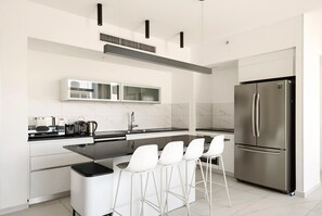 A modern kitchen with a stainless steel refrigerator, a built-in oven, and a bar area with white stools. - Sea&City at Geula with MAMAD by HolyGuest (Tel Aviv)
