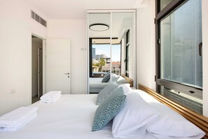 A hotel room with a bed, a large window with blinds, a door, and a view of the cityscape. - Sea&City at Geula with MAMAD by HolyGuest (Tel Aviv)