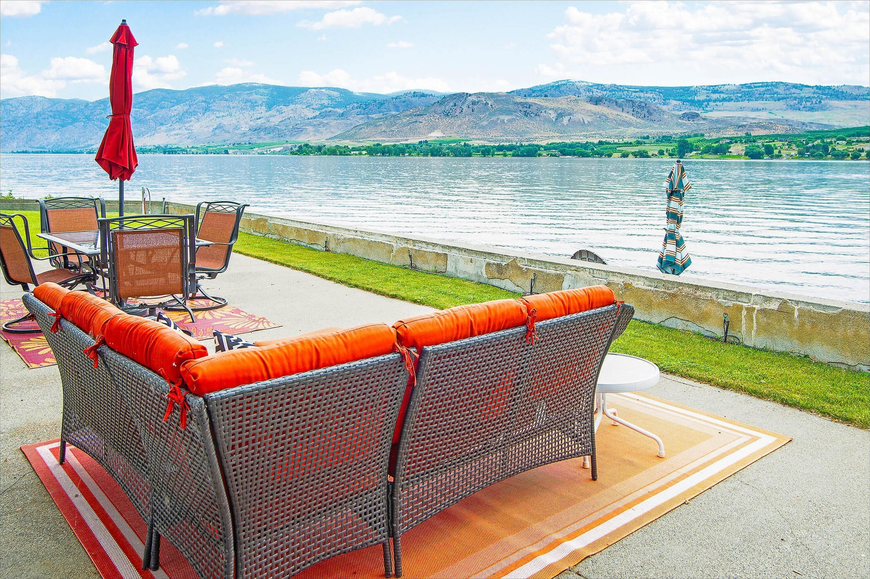 An outdoor seating area with wicker furniture and a red umbrella, overlooking a lake with mountains in the background.