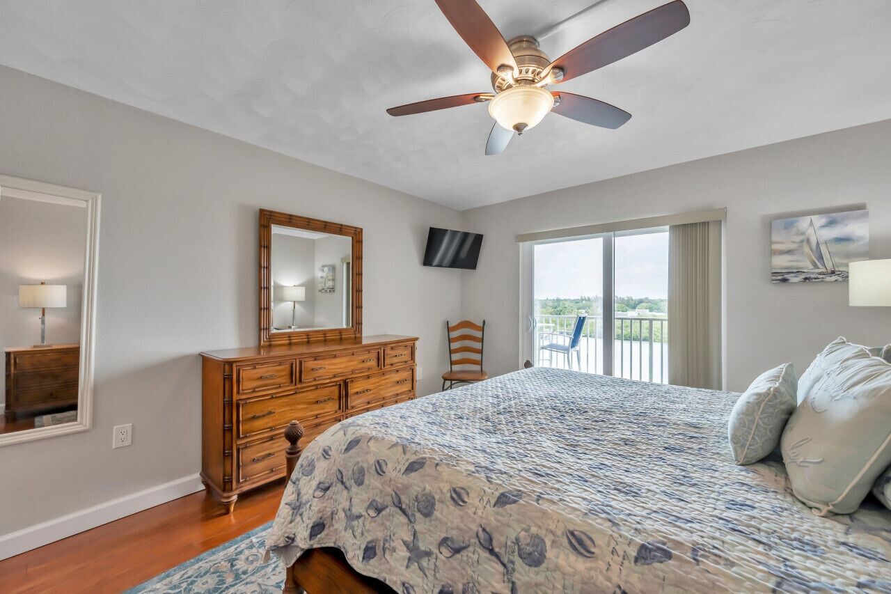 A bedroom with a bed, wooden dresser, ceiling fan, large window, and a balcony.