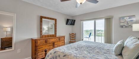 A bedroom with a bed, wooden dresser, ceiling fan, large window, and a balcony.