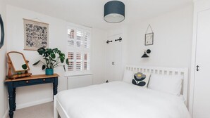 Et soveværelse med en seng, et natbord med et spejl, en potteplante, et vindue med persienner, en vægmonteret lampe og en radiator.