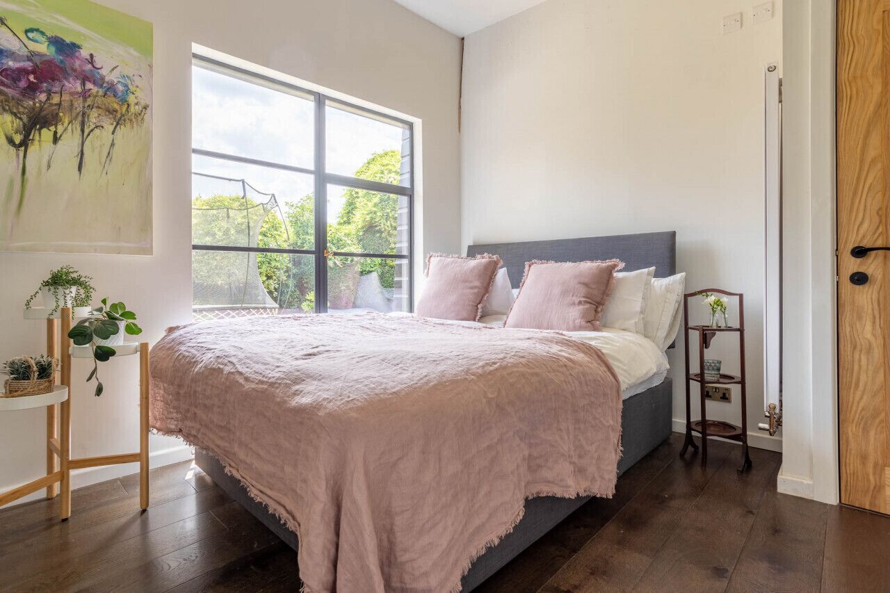 Un dormitorio con un ventanal grande, una cama con ropa de cama rosa, una mesita de noche pequeña con plantas y una puerta de madera.