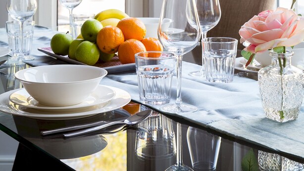 Una mesa de comedor dispuesta con platos, tazones, cubiertos, vasos y un florero con una flor.