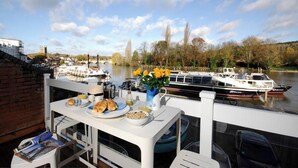 Une table à manger avec des aliments pour le petit-déjeuner et un vase de fleurs jaunes sur un balcon donnant sur une rivière, avec des bateaux et un pont en arrière-plan.