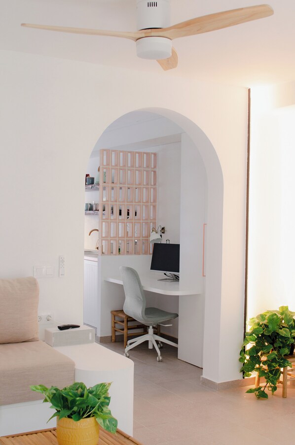 Un espace de bureau moderne et minimaliste, doté d’un bureau aux lignes courbes, d’un ventilateur de plafond et d’un coin salon confortable agrémenté d’une plante.