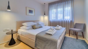 Een net opgemaakt bed met kussens, een nachtkastje met een lamp, een stoel en een raam met gordijnen.