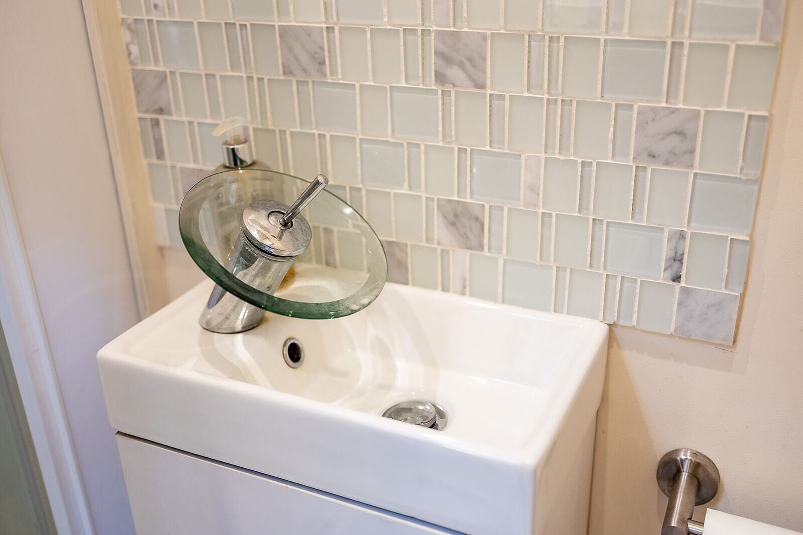 Un baño con lavamanos blanco, dispensador de jabón de vidrio y salpicadero de azulejos.