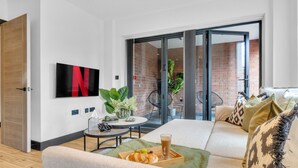 A modern living room with a sofa, a coffee table with a tray of croissants and a drink, a TV mounted on the wall, and a sliding glass door leading to an outdoor area with a brick wall and plants. - homely - Luxury Apartments Finchley (London)