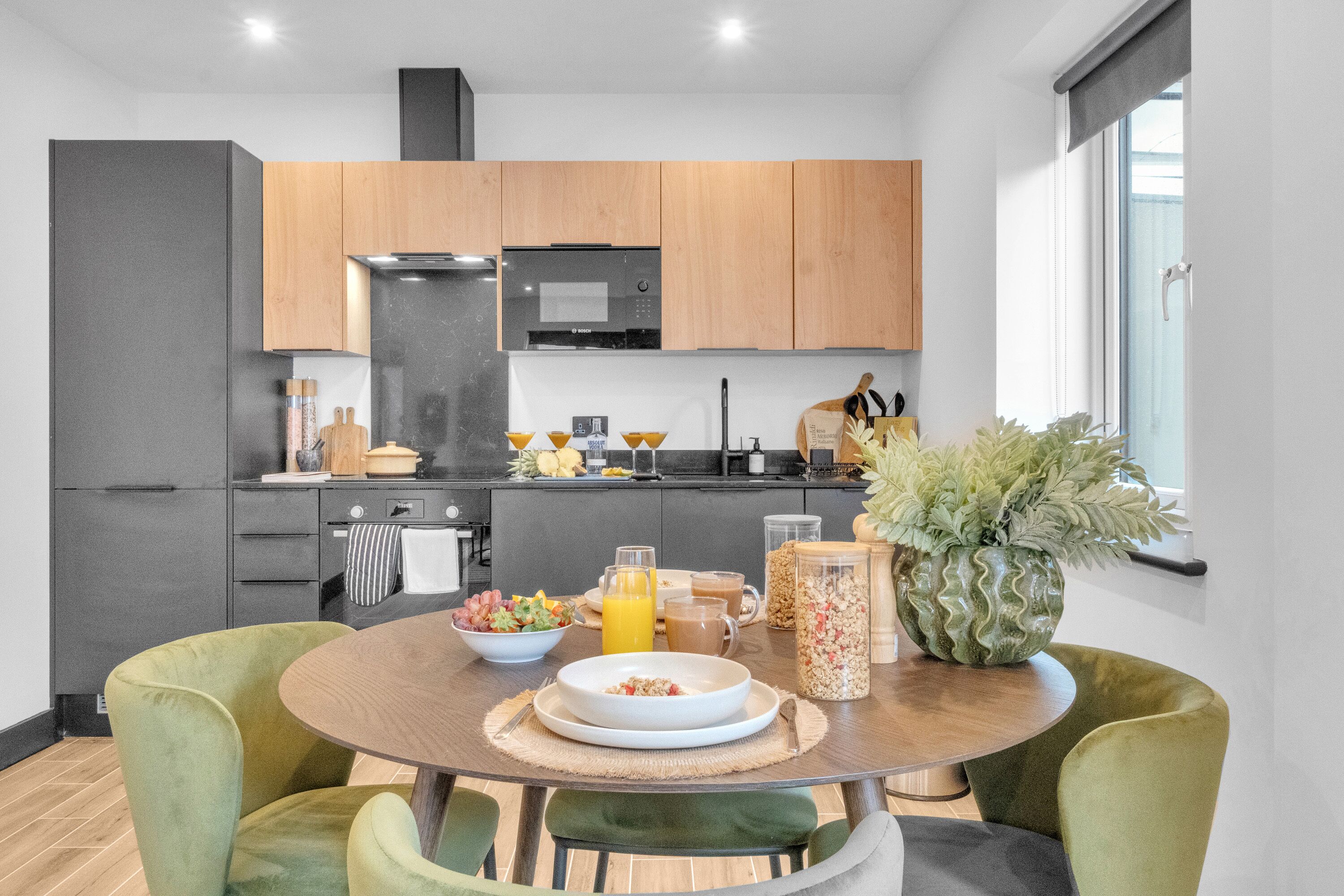 Una cocina moderna con armarios de madera, horno empotrado y comedor con mesa redonda y sillas verdes.
