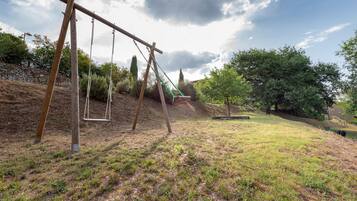 A playground with swings and a slide, surrounded by trees and a stone wall.