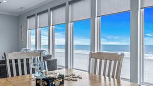 A room with a large window overlooking a beach, a wooden table with a glass of water and dominoes, and a chair. - Southern Charm: Oceanfront w/Pool Near Garden City Pier (Murrells Inlet)