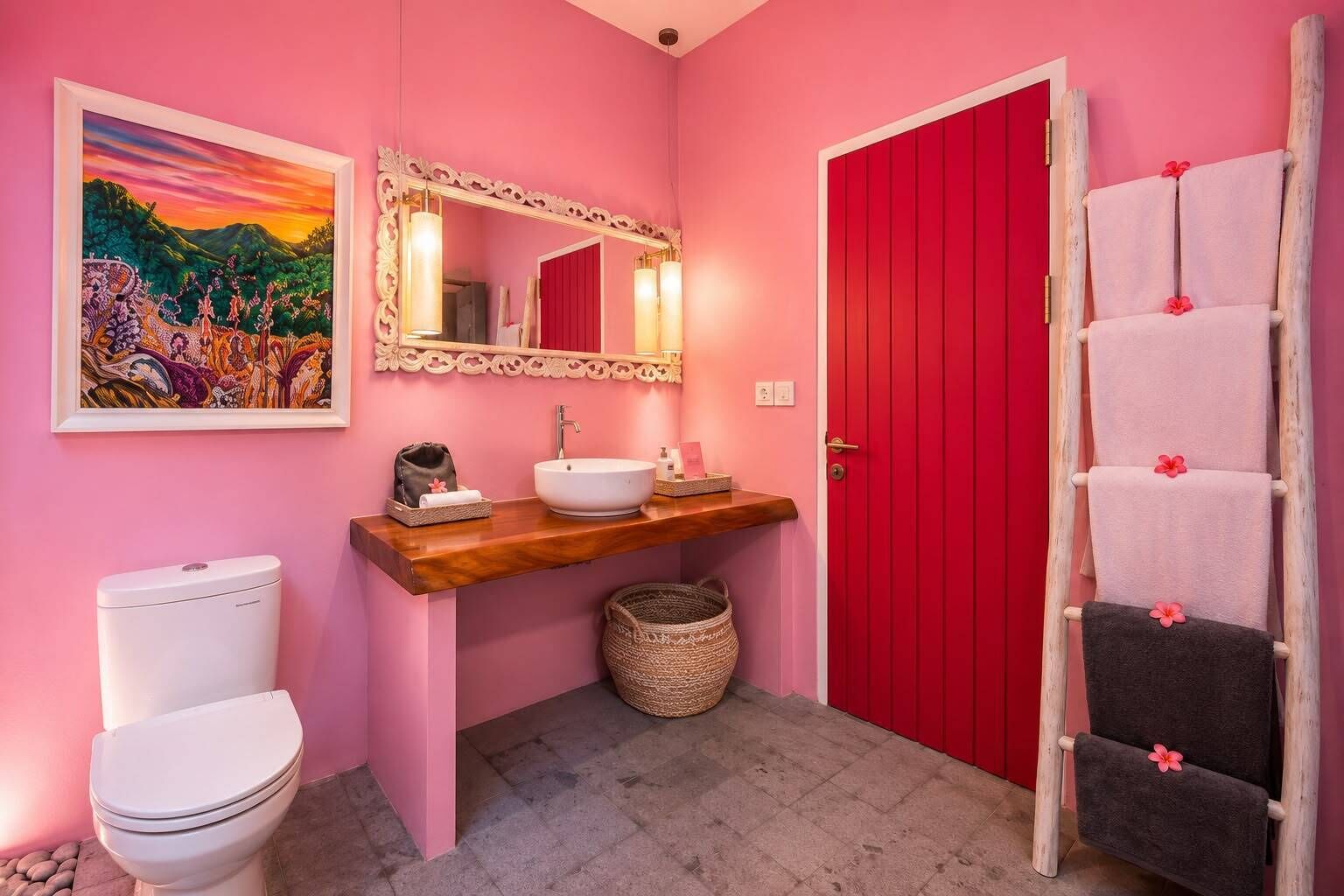 Une salle de bain avec un mur rose, un meuble-lavabo en bois, un lavabo blanc, des toilettes, une porte rouge, un miroir avec un cadre décoratif et un porte-serviettes avec des serviettes.