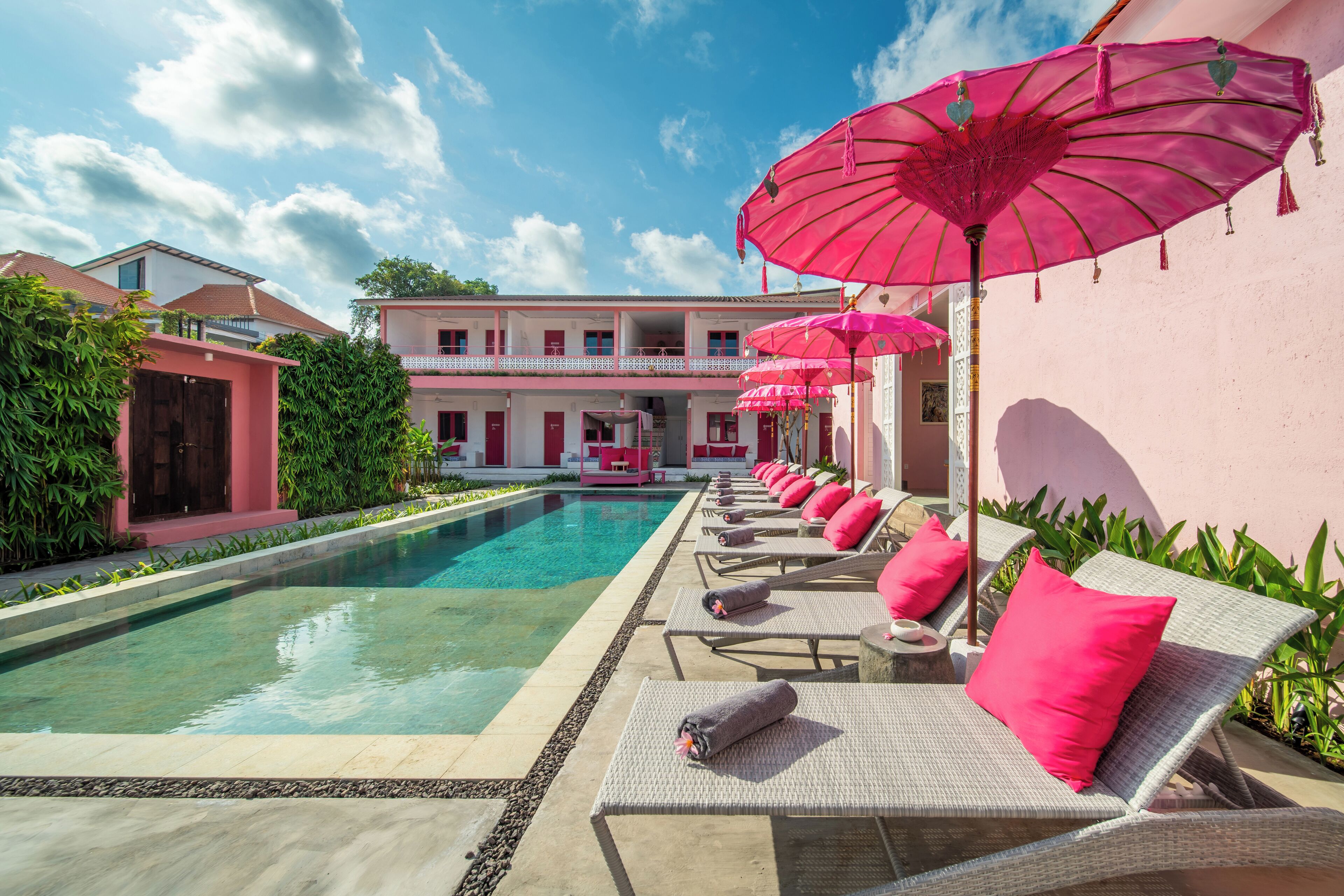 Área ao lado da piscina com espreguiçadeiras e guarda-sóis cor-de-rosa, uma piscina límpida e um edifício com varandas.
