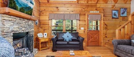 A cozy log cabin interior with a stone fireplace, a black leather sofa, a wooden coffee table, and a large painting of a mountain landscape.