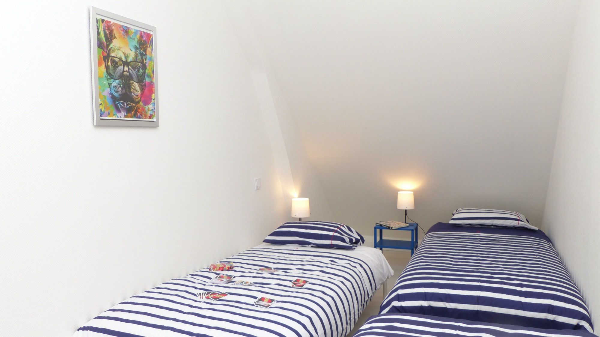 A small, single-room accommodation with two beds, each with striped bedding and a pillow. There is a colorful painting on the wall and a small table with a lamp between the beds.