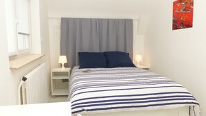 A small, neatly arranged bedroom with a bed, two bedside lamps, a window with a curtain, and a radiator.