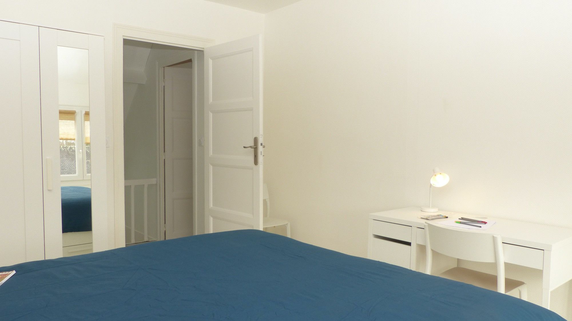 Une chambre à coucher avec un lit, un bureau et une chaise. Une porte donne accès à une autre pièce.