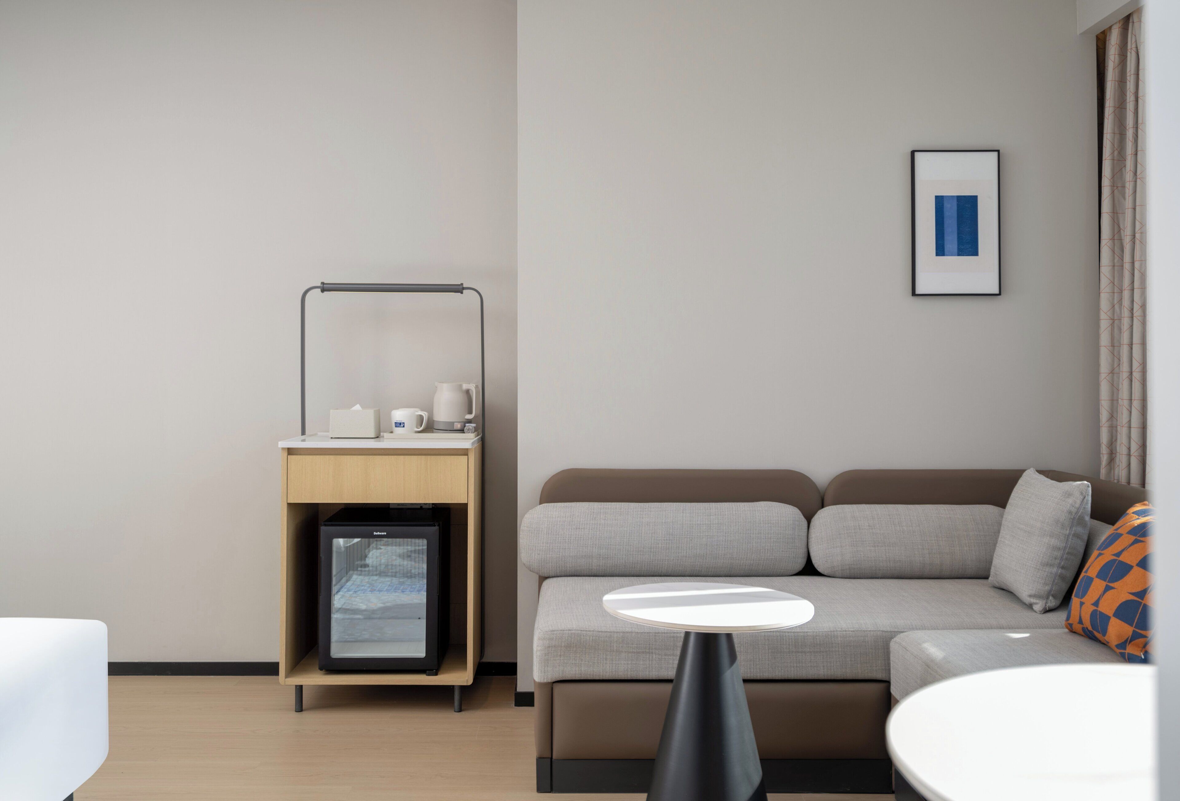 A modern hotel room with a sofa, a small table, a minibar, and a framed picture on the wall.