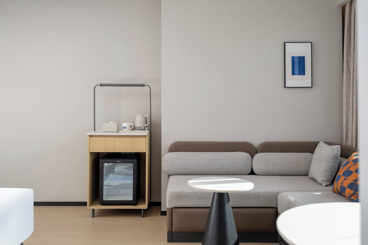 Habitación de hotel moderna con sofá, una mesita, un minibar y un cuadro enmarcado en la pared.